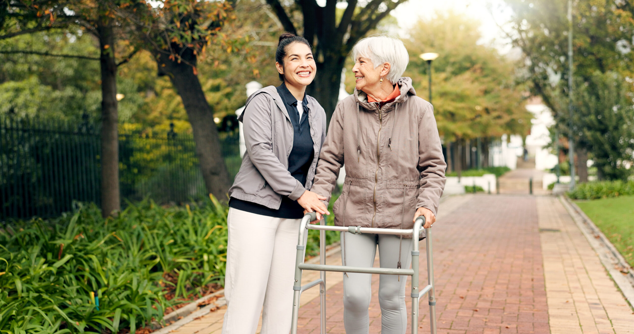 Aging woman (right) walks through a park with the support of a walker and her caregiver.